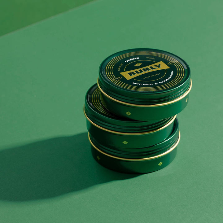 Stack of 3 Burly Creme pomade tins on a green background
